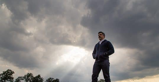 A man in a hoodie standing in a field under a cloudy sky.