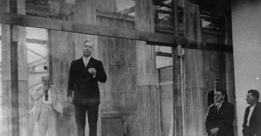 A group of men standing in front of a building, poised for public speaking.