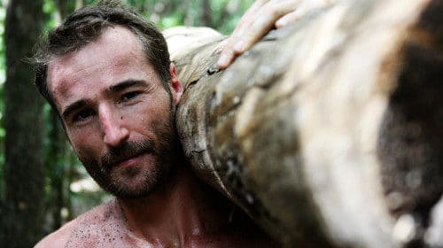 A shirtless man carrying a log in the woods.
