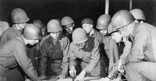 During WWII, a group of soldiers gathers around to look at a piece of paper.
