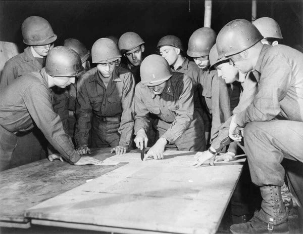 During WWII, a group of soldiers gathers around to look at a piece of paper.