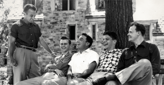 Several casual men are sitting on a bench in front of a building.