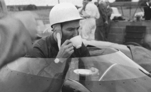 A man in a helmet sipping tea from a cup in a race car, honoring a manly tradition.