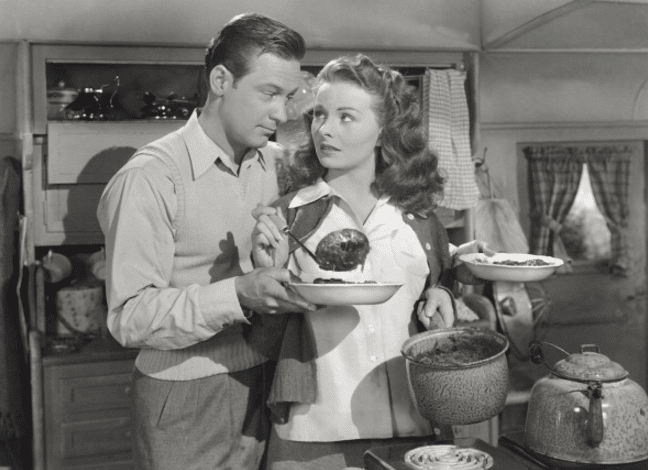 Vintage woman pouring food in the plate of the man in the kitchen.