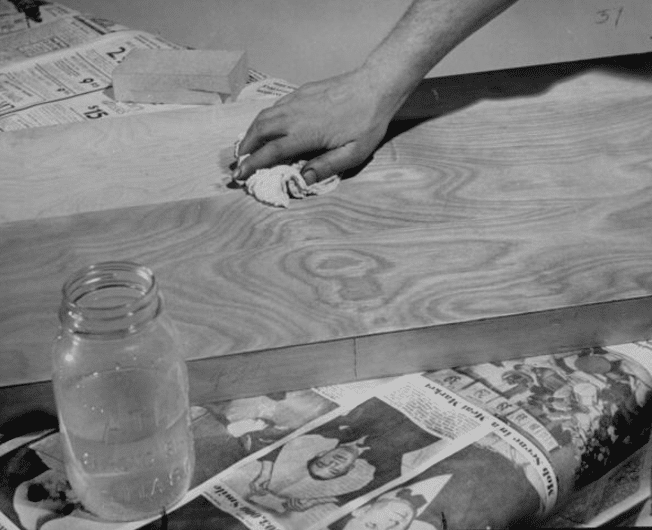 Vintage man rubbing finish on a wood board while having finish solution in a glass jar.