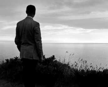 A man in a suit is standing on a hill overlooking the ocean, deep in reflection.
