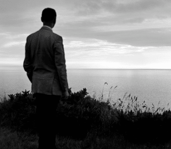A man in a suit is standing on a hill overlooking the ocean, deep in reflection.