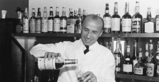 A man pouring bourbon in front of a liquor cabinet.