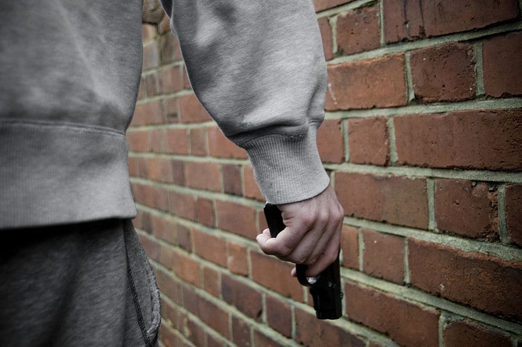 A man brandishing a gun in front of a brick wall, portraying social aggression.