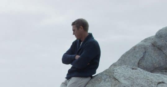 A man wearing a cardigan sweater is sitting on top of a large rock.
