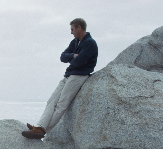 A man wearing a cardigan sweater is sitting on top of a large rock.