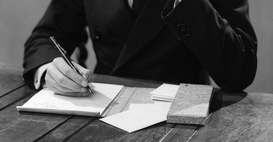 A man in a suit writing on a notebook, composing a letter.