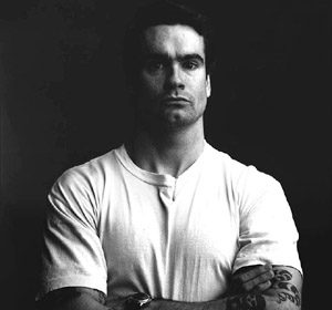 A black and white photo of a man with tattoos, reminiscent of Henry Rollins.