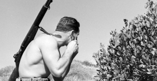 A man with a rifle in front of a bush showing fidelity to his surroundings.