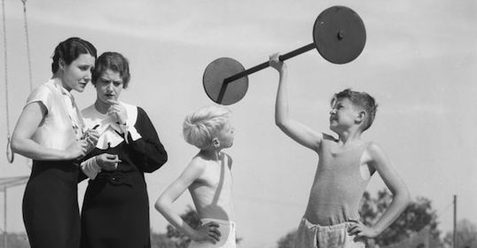 An old black and white photo of a woman lifting weights in front of a group of kids.