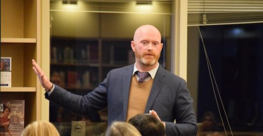 A bald man is giving a presentation in front of a group of people.