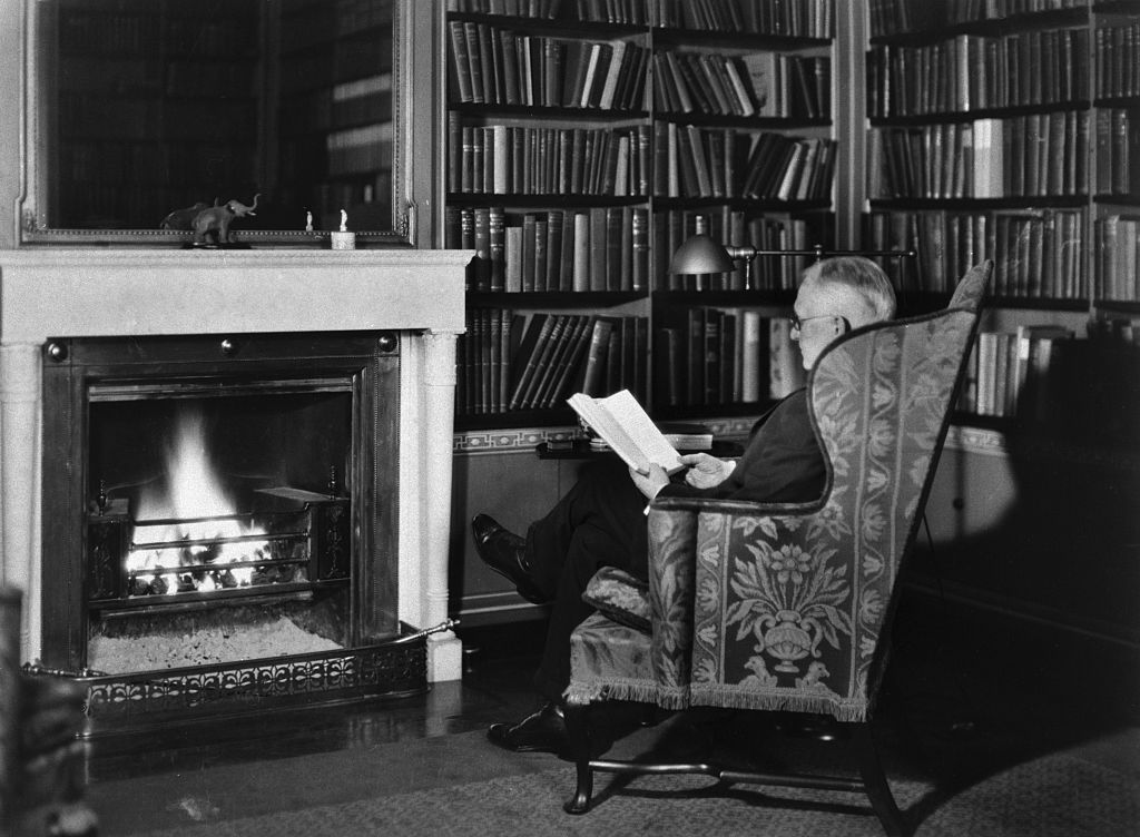 A man reading a book in front of a fireplace.