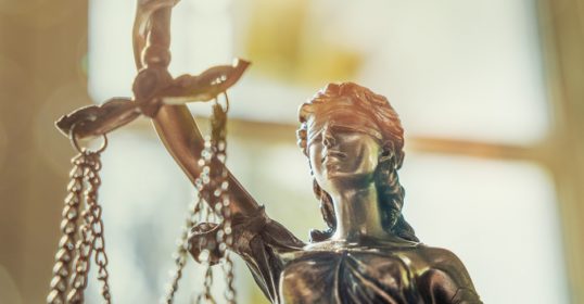 A manvotional statue of justice sitting with scales in front of a window.