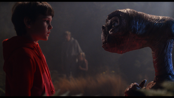 Scene from the movie "E.T" in which a boy is talking to an alien and person is standing in background.