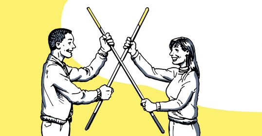 A man and his partner stand holding sticks in front of a yellow background.