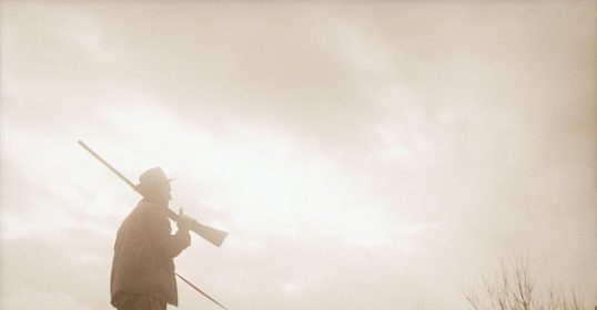 A man with a rifle holding a leash, walking his dog.