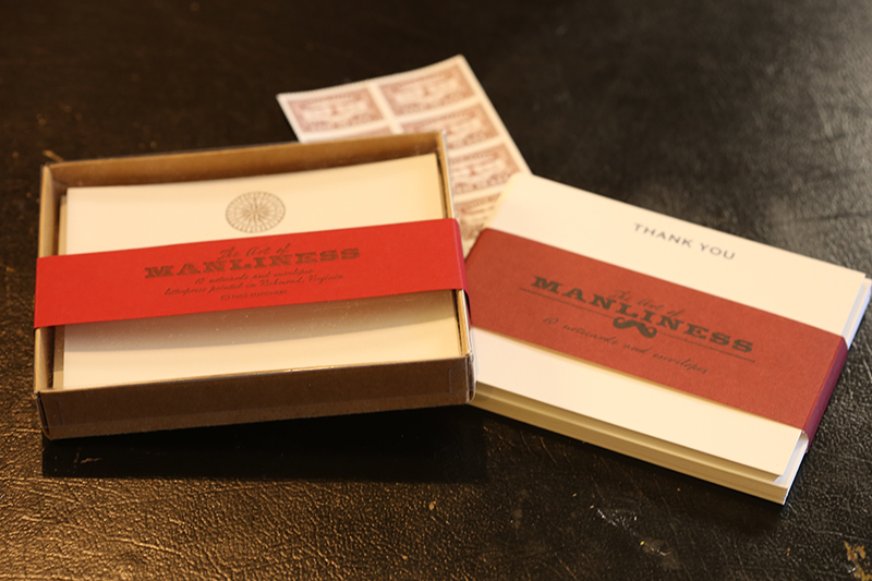 An open box displaying a set of stationery labelled "the art of manliness" with a thank you card placed alongside it on a dark textured surface.