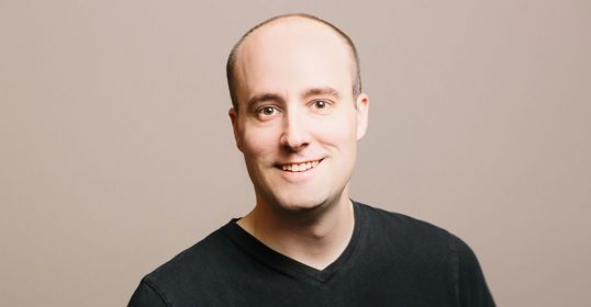 A bald man in a black v-neck shirt smiles while discussing mental models on his podcast.
