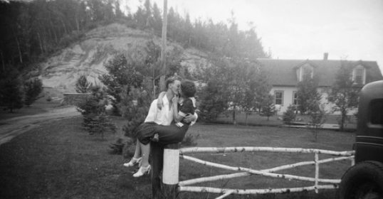 A newly married woman is leaning on a fence in front of a house, offering advice to passersby.