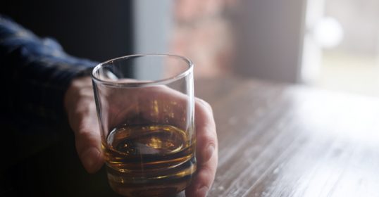 Enhance the image of a man enjoying whiskey, holding a glass on a wooden table.