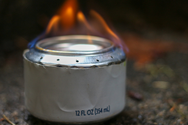 A soda can stove siting on the ground with a flame.