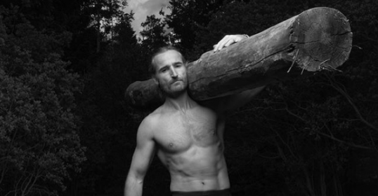 A shirtless man carrying a log through the woods.