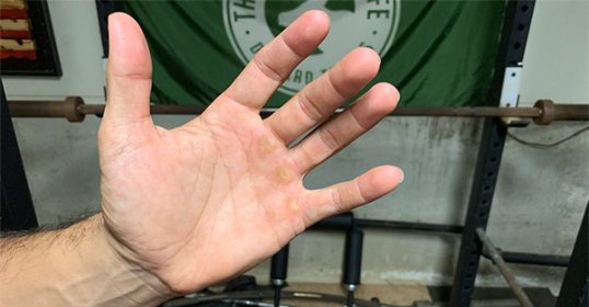 A man's hand in front of a heavy barbell in the gym, showing signs of calluses from weightlifting.