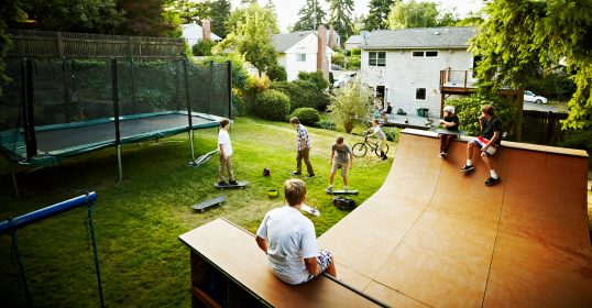 A group of kids create playing on a skateboard ramp in a neighborhood backyard.