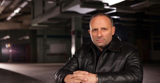 A man in a leather jacket sits on a chair in an empty building, possibly recording a new podcast episode.