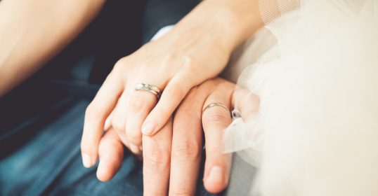 A bride and groom's hands strengthen each other's wedding rings.
