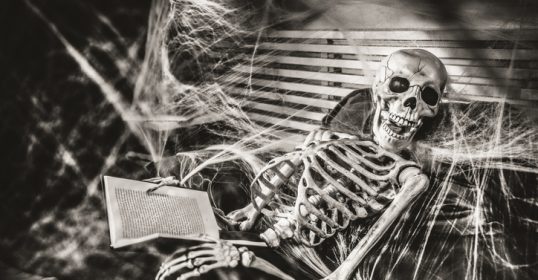 A classic skeleton sits in a chair, reading a book.