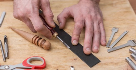 A man is making a leather belt with a pair of scissors on a wooden table.