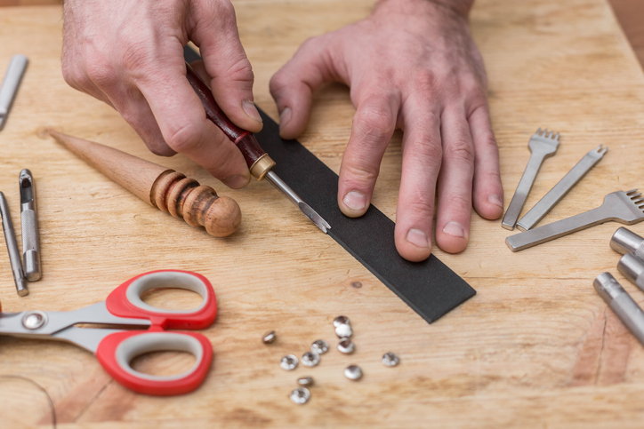 A man is making a leather belt with a pair of scissors on a wooden table.