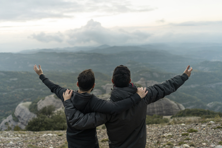 Two friends standing on top of a mountain, arms outstretched.