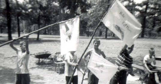 An untold story of a group of boys holding flags in a dirt field during the Robbers Cave Experiment.