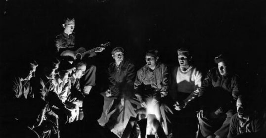 A group of men sitting around a campfire, enjoying the lost pleasures of group singing.
