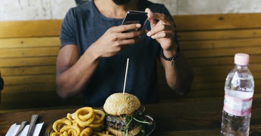A man is still taking a picture of a burger on his phone.