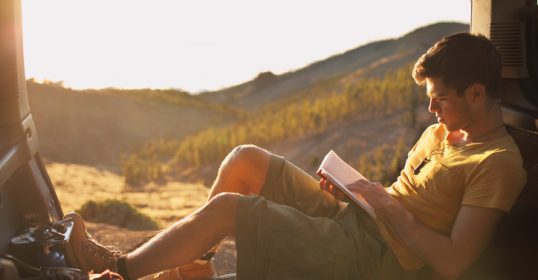 A man sitting in the back seat of a car reading for pleasure.