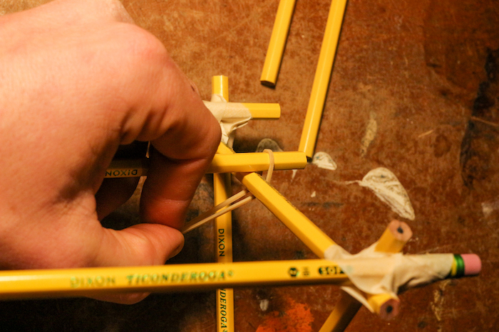 Attaching a pencil halfway up one side of the supports using rubber bands.