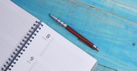 During quarantine, maintaining sanity can be a challenge. Writing in a notebook with a pen on a blue wooden table can be a helpful outlet.