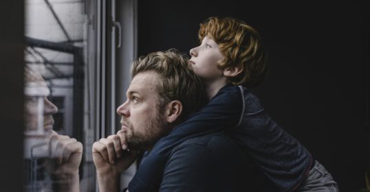 A man and his son looking out a window during the pandemic.