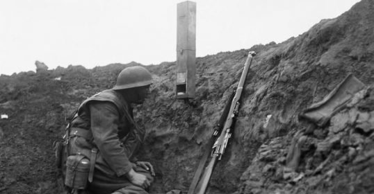 A soldier kneeling in a trench with a rifle uses a mirror periscope designed by kids for better visibility.