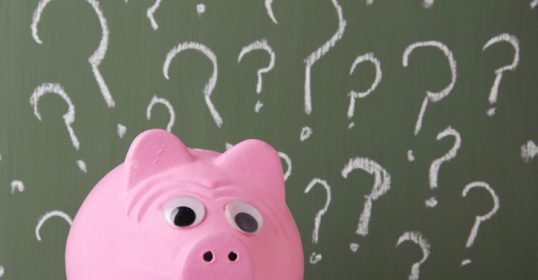 A pink piggy bank sits in front of a chalkboard covered in question marks.