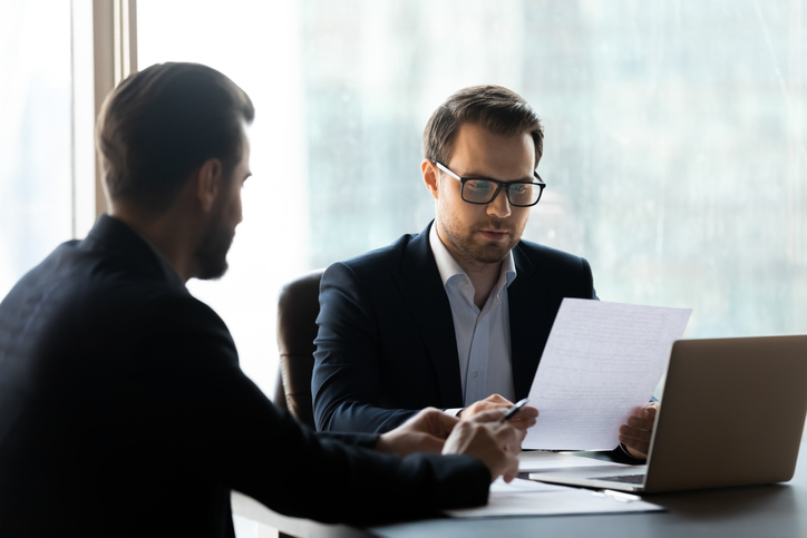 Two men are in a business meeting and one of them is holding a paper with a laptop in front of him.