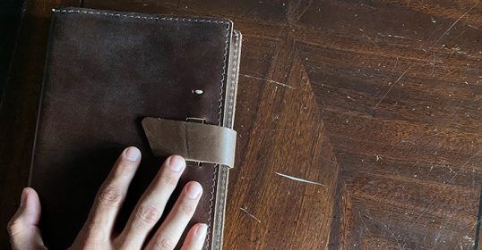 A person's hand holding a brown notebook on a wooden table, showcasing their love for journaling.